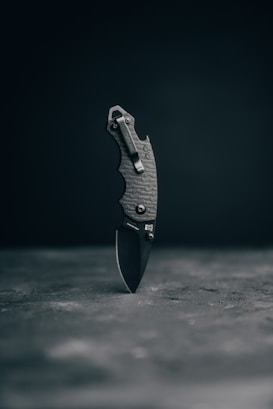 A folded pocket knife with a textured grip is balanced vertically on its end against a dark, neutral background. The surface beneath the knife is slightly rough and muted.