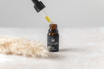A small dark glass bottle with a label sits on a light surface, featuring a logo and text indicating it is CBD oil. Above the bottle, a dropper is held, releasing a drop of oil back into the bottle. To the side, there is a soft-focus image of beige, fluffy pampas grass.