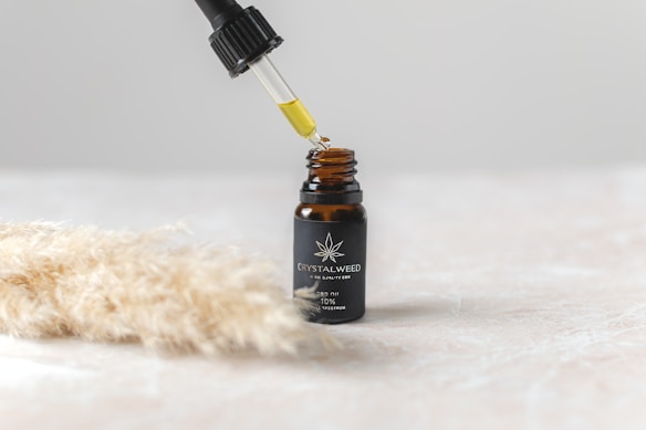 A small dark glass bottle with a label sits on a light surface, featuring a logo and text indicating it is CBD oil. Above the bottle, a dropper is held, releasing a drop of oil back into the bottle. To the side, there is a soft-focus image of beige, fluffy pampas grass.