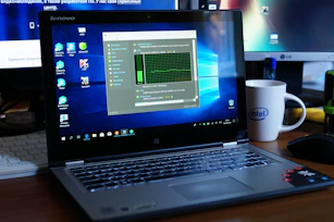 A Lenovo laptop displayed on a wooden desk with a screen showing a system performance window and several desktop icons. Behind it, there are additional computer monitors, one of which has a visible LG logo, and a white coffee mug with an Intel logo. Various office items like a mouse and a keyboard can also be seen.