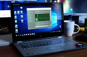 A Lenovo laptop displayed on a wooden desk with a screen showing a system performance window and several desktop icons. Behind it, there are additional computer monitors, one of which has a visible LG logo, and a white coffee mug with an Intel logo. Various office items like a mouse and a keyboard can also be seen.