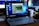 A Lenovo laptop displayed on a wooden desk with a screen showing a system performance window and several desktop icons. Behind it, there are additional computer monitors, one of which has a visible LG logo, and a white coffee mug with an Intel logo. Various office items like a mouse and a keyboard can also be seen.