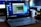 A Lenovo laptop displayed on a wooden desk with a screen showing a system performance window and several desktop icons. Behind it, there are additional computer monitors, one of which has a visible LG logo, and a white coffee mug with an Intel logo. Various office items like a mouse and a keyboard can also be seen.