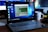 A Lenovo laptop displayed on a wooden desk with a screen showing a system performance window and several desktop icons. Behind it, there are additional computer monitors, one of which has a visible LG logo, and a white coffee mug with an Intel logo. Various office items like a mouse and a keyboard can also be seen.