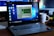 A Lenovo laptop displayed on a wooden desk with a screen showing a system performance window and several desktop icons. Behind it, there are additional computer monitors, one of which has a visible LG logo, and a white coffee mug with an Intel logo. Various office items like a mouse and a keyboard can also be seen.