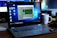 A Lenovo laptop displayed on a wooden desk with a screen showing a system performance window and several desktop icons. Behind it, there are additional computer monitors, one of which has a visible LG logo, and a white coffee mug with an Intel logo. Various office items like a mouse and a keyboard can also be seen.