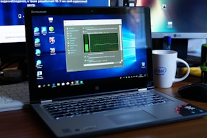 A Lenovo laptop displayed on a wooden desk with a screen showing a system performance window and several desktop icons. Behind it, there are additional computer monitors, one of which has a visible LG logo, and a white coffee mug with an Intel logo. Various office items like a mouse and a keyboard can also be seen.