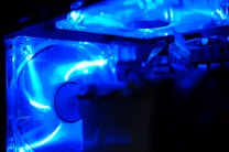 A close-up view of the interior of a computer case, featuring illuminated cooling fans and components. The image is dominated by a vibrant blue light that creates a futuristic and tech-oriented atmosphere.