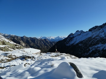 A snowy mountain landscape with a view of rugged peaks under a clear blue sky. The foreground features a snow-covered surface with rocky outcrops, and there is a small structure with a flag positioned centrally among the mountains.