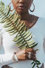 A person in a white top holds a large green fern leaf in front of them. They are accessorized with hoop earrings, a necklace, and several bracelets. Their lips are slightly parted, and a soft shadow falls across their face.