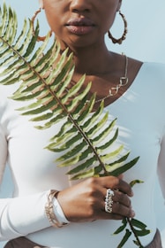 A person in a white top holds a large green fern leaf in front of them. They are accessorized with hoop earrings, a necklace, and several bracelets. Their lips are slightly parted, and a soft shadow falls across their face.