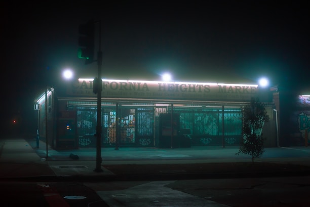 A small market is illuminated by neon lights on a foggy night, creating a moody and atmospheric scene. The store's lights cast a glow on the wet pavement, and the surrounding area appears desolate and quiet. Faint street art can be seen on the walls.