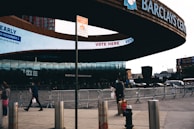 The image depicts the entrance of Barclays Center, with 'Vote Here' signage displayed prominently. Several pedestrians, some wearing masks, are walking past metal barriers set up in front of the building. The urban environment features buildings and a billboard in the background.