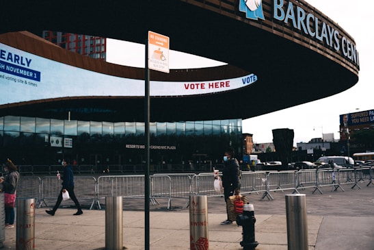 The image depicts the entrance of Barclays Center, with 'Vote Here' signage displayed prominently. Several pedestrians, some wearing masks, are walking past metal barriers set up in front of the building. The urban environment features buildings and a billboard in the background.