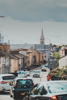 A busy urban street filled with cars and pedestrians. Buildings line both sides of the road, and a church spire rises prominently in the background under an overcast sky.