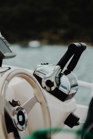 Close view of a boat helm with a white steering wheel and a chrome throttle lever beside it, water blurred beyond the console