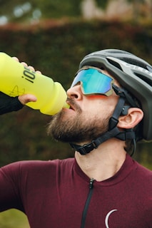 A cyclist is quenching his thirst with a bright yellow water bottle while wearing a burgundy cycling jersey and protective gear. He sports sunglasses with blue reflective lenses and a helmet, with a lush green background.