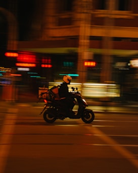 A motorcyclist, wearing a helmet and carrying a delivery box, rides swiftly on a scooter through a dimly lit urban street. The background is blurred, emphasizing the motion and speed of the motorbike.