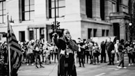 A man holding a microphone is speaking passionately to a crowd gathered on a city street. The scene is monochrome, with an American flag visible on the left. People in the background are holding signs, capturing the essence of a protest or public demonstration.