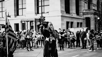 A man holding a microphone is speaking passionately to a crowd gathered on a city street. The scene is monochrome, with an American flag visible on the left. People in the background are holding signs, capturing the essence of a protest or public demonstration.