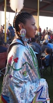 A person dressed in vibrant traditional attire, adorned with intricate beadwork and feather accessories. The background has a crowd of people, suggesting a cultural event.