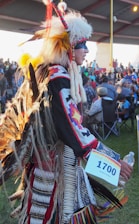 A person dressed in traditional Indigenous attire, featuring a headdress with feathers and a detailed, colorful outfit. They are holding a numbered card and a water bottle, participating in an event with an audience visible in the background under a canopy.