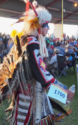A person dressed in traditional Indigenous attire, featuring a headdress with feathers and a detailed, colorful outfit. They are holding a numbered card and a water bottle, participating in an event with an audience visible in the background under a canopy.