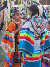 Two individuals in brightly colored, traditional attire engaged in conversation. Their garments feature vibrant stripes and intricate beadwork with a variety of colors including orange, blue, and red. The setting appears to be a cultural event with others in the background wearing similar attire adorned with feathers.