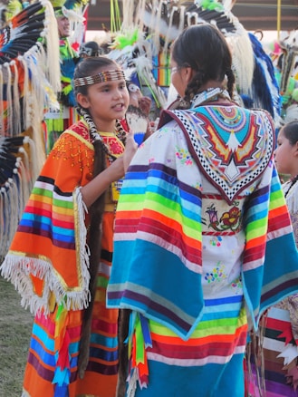 Two individuals in brightly colored, traditional attire engaged in conversation. Their garments feature vibrant stripes and intricate beadwork with a variety of colors including orange, blue, and red. The setting appears to be a cultural event with others in the background wearing similar attire adorned with feathers.
