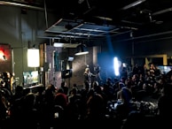A film or video production set with a large group of people, featuring lights, cameras, and equipment. A person in the center is managing the setup, surrounded by an audience seated and observing the activity. The room appears dark with key lighting focused on the set, creating a dramatic atmosphere. Screens and monitors display content related to the production process.