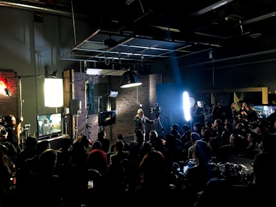 A film or video production set with a large group of people, featuring lights, cameras, and equipment. A person in the center is managing the setup, surrounded by an audience seated and observing the activity. The room appears dark with key lighting focused on the set, creating a dramatic atmosphere. Screens and monitors display content related to the production process.