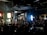 A film or video production set with a large group of people, featuring lights, cameras, and equipment. A person in the center is managing the setup, surrounded by an audience seated and observing the activity. The room appears dark with key lighting focused on the set, creating a dramatic atmosphere. Screens and monitors display content related to the production process.