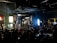 A film or video production set with a large group of people, featuring lights, cameras, and equipment. A person in the center is managing the setup, surrounded by an audience seated and observing the activity. The room appears dark with key lighting focused on the set, creating a dramatic atmosphere. Screens and monitors display content related to the production process.
