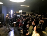 A person appears to be giving a presentation or demonstration in front of a seated audience in a dimly lit room. The room has professional lighting equipment, suggesting a workshop or seminar in the field of photography or film. Attendees are attentively watching, some taking notes.