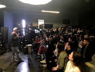 A person appears to be giving a presentation or demonstration in front of a seated audience in a dimly lit room. The room has professional lighting equipment, suggesting a workshop or seminar in the field of photography or film. Attendees are attentively watching, some taking notes.