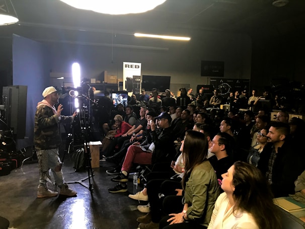 A person appears to be giving a presentation or demonstration in front of a seated audience in a dimly lit room. The room has professional lighting equipment, suggesting a workshop or seminar in the field of photography or film. Attendees are attentively watching, some taking notes.