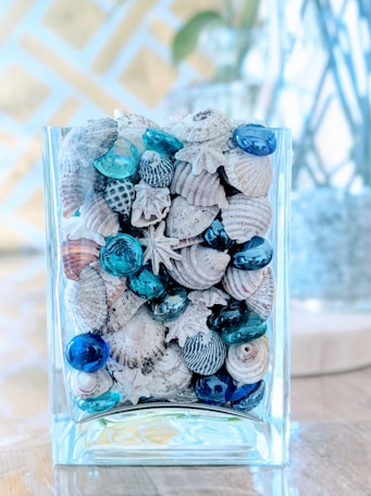 A glass container filled with various seashells and turquoise glass pebbles is placed on a wooden surface. The seashells vary in size and shape, including spiral, scallop, and conical types, with natural earthy tones. The glass pebbles interspersed among the shells add a vibrant blue hue, enhancing the oceanic theme. The background is softly blurred, with floral designs creating a tranquil atmosphere.