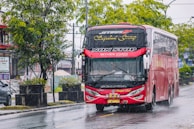 A large red bus driving on a wet road with rain visible around it. The bus has 'Non Stop Skyview Coach' written on the front and is a Scania brand vehicle. Trees and greenery are on the side of the road, with a car partially visible in the background. The scene suggests a rainy day in an urban setting.