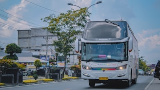 A large, modern bus with a sleek design is traveling down an urban street lined with trees and commercial buildings. The bus features prominent branding and a license plate, while the surrounding area shows power lines and street signage under a clear sky.