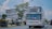 A large, modern bus with a sleek design is traveling down an urban street lined with trees and commercial buildings. The bus features prominent branding and a license plate, while the surrounding area shows power lines and street signage under a clear sky.