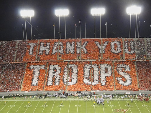 A football stadium packed with fans holding colored cards to create a large display that reads 'THANK YOU TROOPS.' The display covers an entire section of the stadium seats and is illuminated by bright stadium lights. Players and staff are visible on the field below.