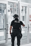 A police officer is standing with their back facing the camera, positioned near a set of double-glass doors. The officer is dressed in tactical gear which includes a helmet, a communication device attached to the shoulder, and utility belts with various tools. The scene appears to be indoors with bright lighting and the reflection of sunlight on the glass.