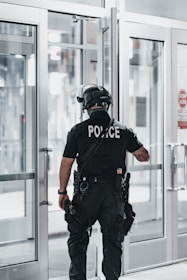 A police officer is standing with their back facing the camera, positioned near a set of double-glass doors. The officer is dressed in tactical gear which includes a helmet, a communication device attached to the shoulder, and utility belts with various tools. The scene appears to be indoors with bright lighting and the reflection of sunlight on the glass.
