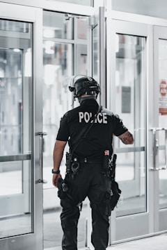 A police officer is standing with their back facing the camera, positioned near a set of double-glass doors. The officer is dressed in tactical gear which includes a helmet, a communication device attached to the shoulder, and utility belts with various tools. The scene appears to be indoors with bright lighting and the reflection of sunlight on the glass.