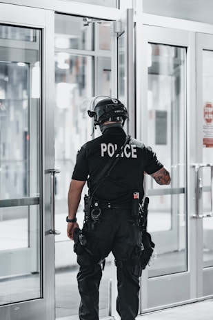 A police officer is standing with their back facing the camera, positioned near a set of double-glass doors. The officer is dressed in tactical gear which includes a helmet, a communication device attached to the shoulder, and utility belts with various tools. The scene appears to be indoors with bright lighting and the reflection of sunlight on the glass.