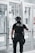 A police officer is standing with their back facing the camera, positioned near a set of double-glass doors. The officer is dressed in tactical gear which includes a helmet, a communication device attached to the shoulder, and utility belts with various tools. The scene appears to be indoors with bright lighting and the reflection of sunlight on the glass.