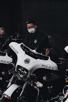 A person wearing a mask is standing next to a row of police motorcycles with 'Denver Police' written on them. The lighting is dim, creating a serious and focused atmosphere.