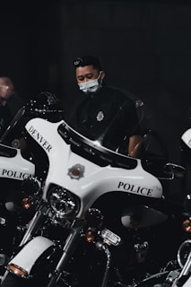 A person wearing a mask is standing next to a row of police motorcycles with 'Denver Police' written on them. The lighting is dim, creating a serious and focused atmosphere.