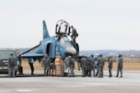 A group of military personnel in uniform are gathered around a fighter jet on an airfield. Some personnel are on the ground, while one person is on top of the aircraft. The jet is a dark blue color with various markings. A vehicle is parked nearby, and the area has an open, expansive feel with a distant tree line.