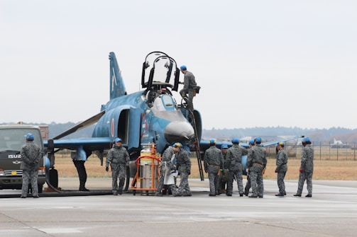 A group of military personnel in uniform are gathered around a fighter jet on an airfield. Some personnel are on the ground, while one person is on top of the aircraft. The jet is a dark blue color with various markings. A vehicle is parked nearby, and the area has an open, expansive feel with a distant tree line.